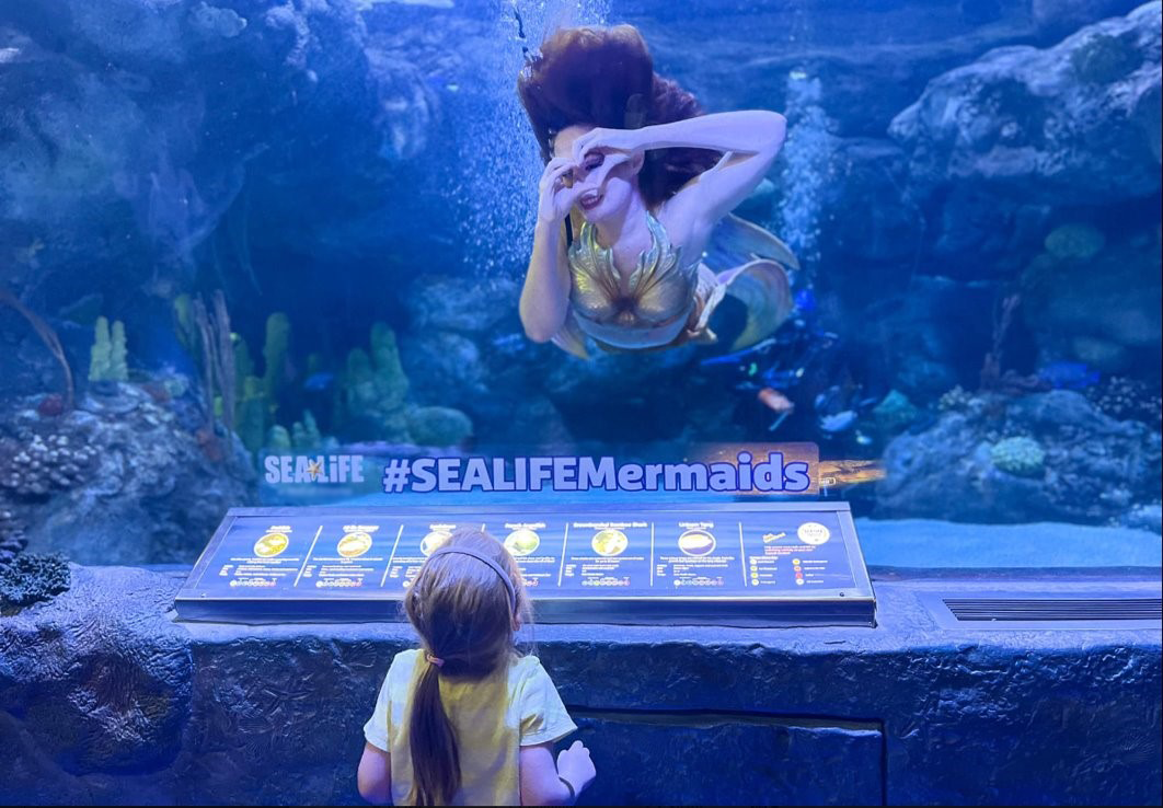 Mermaid underwater making a heart shape with her hands to a child watching in the aquarium