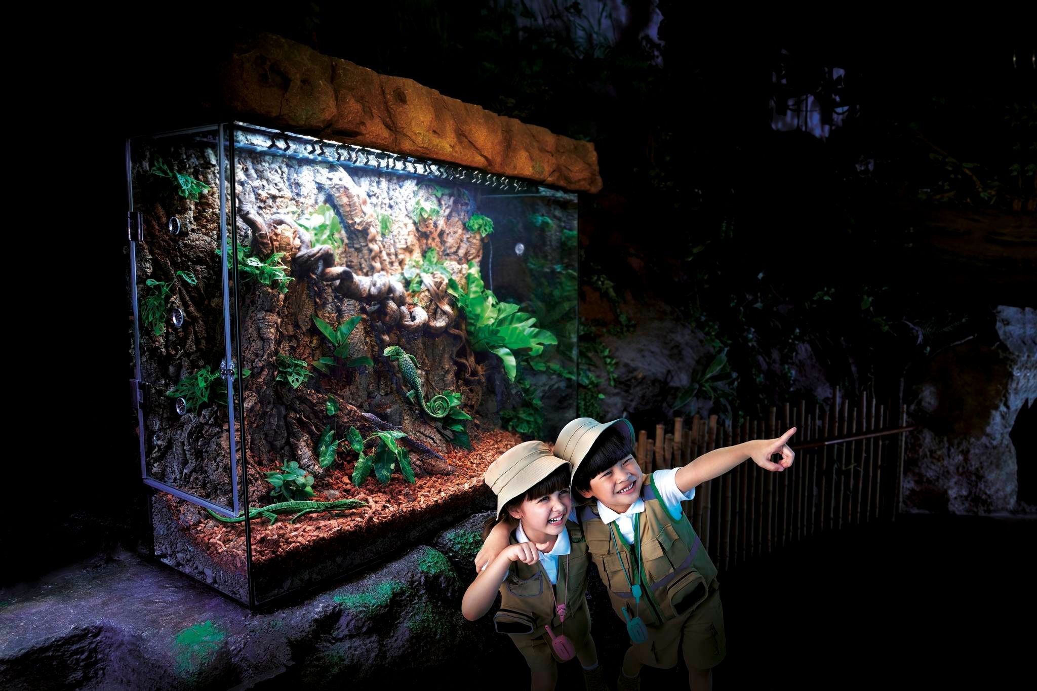 Two boys dressed in safari outfits standing in the lush and vibrant rainforest environment at SEA LIFE Bangkok Ocean World, with diverse plant life and habitat for various creatures.