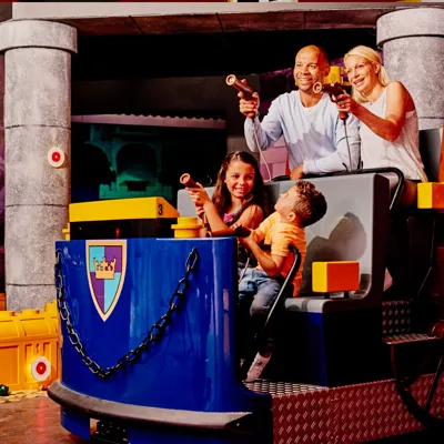 A smiling family of four enjoys an interactive ride at LEGOLAND Discovery Center Dallas-Ft Worth, aiming laser blasters at targets in a medieval-themed setting. The ride vehicle is blue with a LEGO crest, and stone-like columns surround the scene.