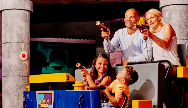 A smiling family of four enjoys an interactive ride at LEGOLAND Discovery Center Dallas-Ft Worth, aiming laser blasters at targets in a medieval-themed setting. The ride vehicle is blue with a LEGO crest, and stone-like columns surround the scene.