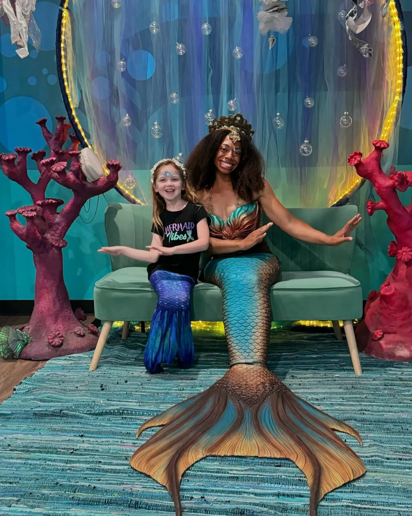 A young girl and a mermaid pose together during the Mermaid Makeover experience at SEA LIFE Grapevine, seated in a colorful underwater-themed photo area. Photo provided by Instagram user @siwanatorwaves.