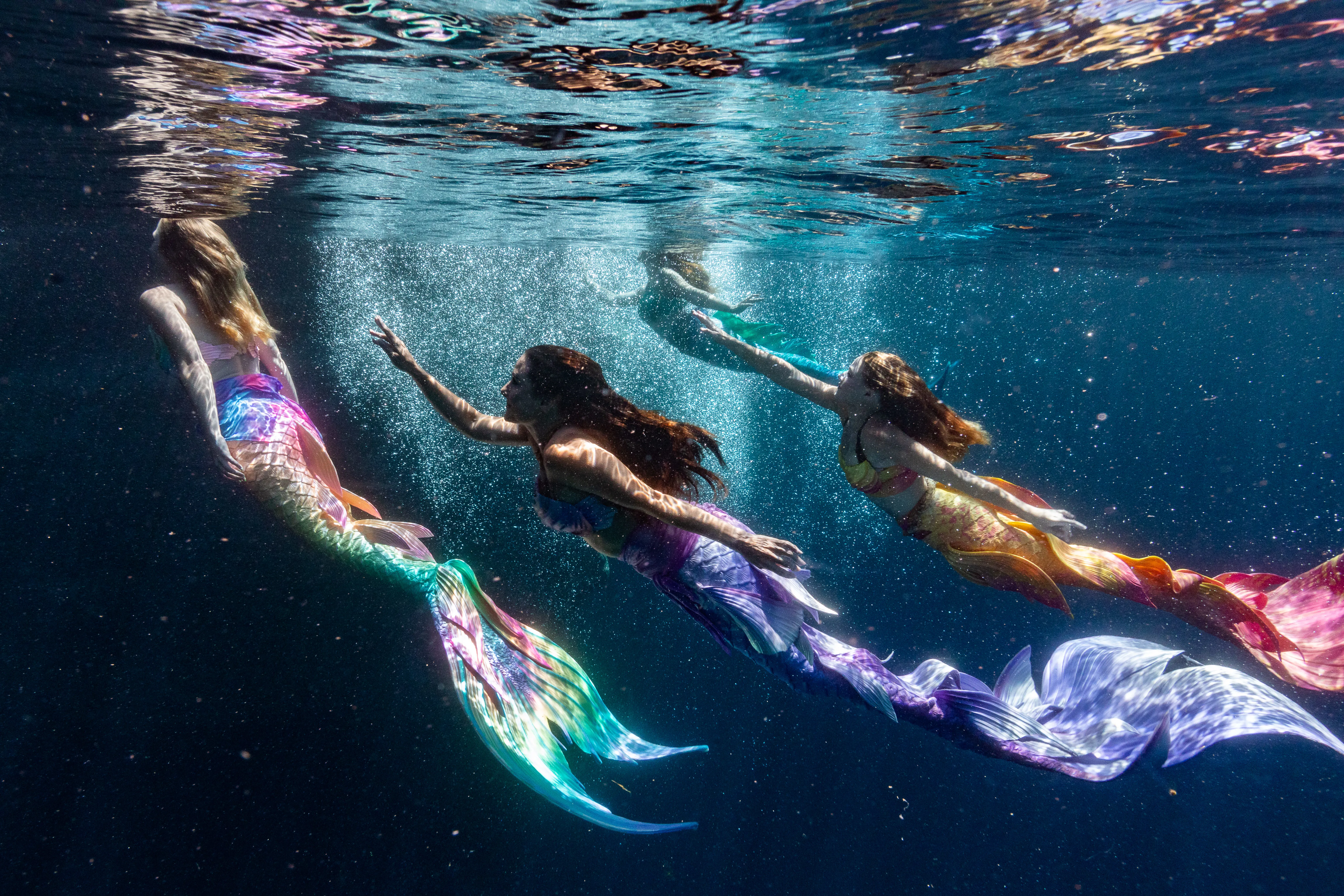 Three mermaids swimming towards the surface of the ocean with a sunbeam streaming down onto their faces.