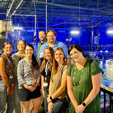 A group of 8 adults posing together in front of the tops of aquatic habitats at SEA LIFE Orlando Aquarium during a behind-the-scenes tour. The group smiles and stands close together, with aquarium tanks and equipment visible in the background, highlighting the exclusive experience of the tour. Image provided by IG user : Thekaitlynelam